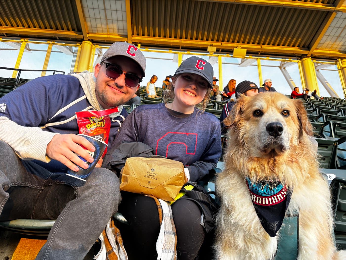 Ty, his girlfriend, and Bo the dog wearing a Guardians bandana in the stands at a Cleveland Guardians game